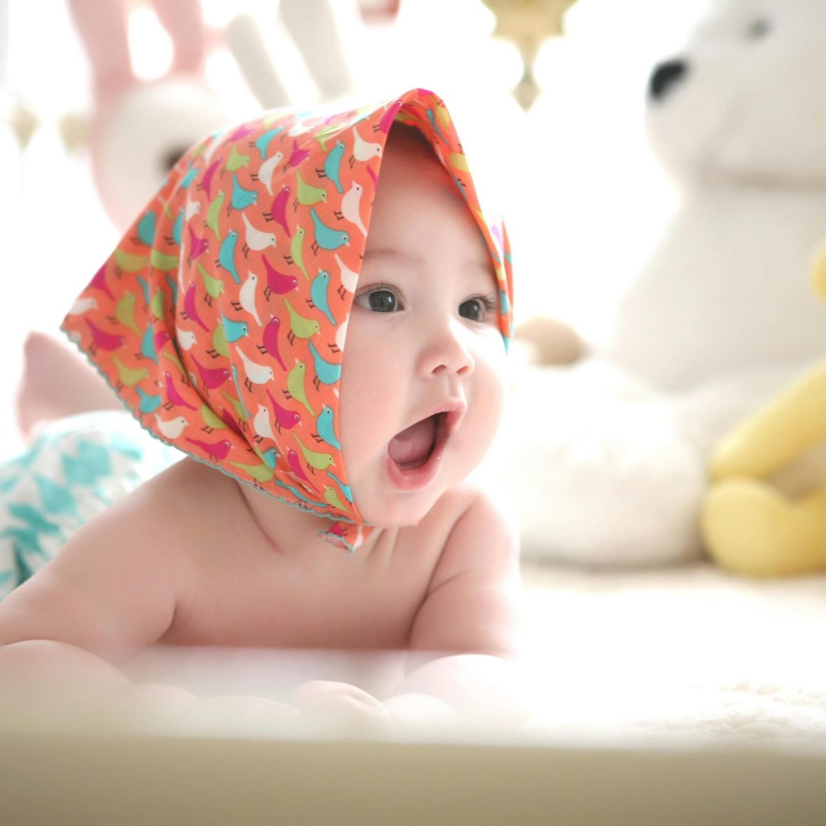 Charming baby with colorful scarf surrounded by cute plush toys, highlighting innocence indoors.